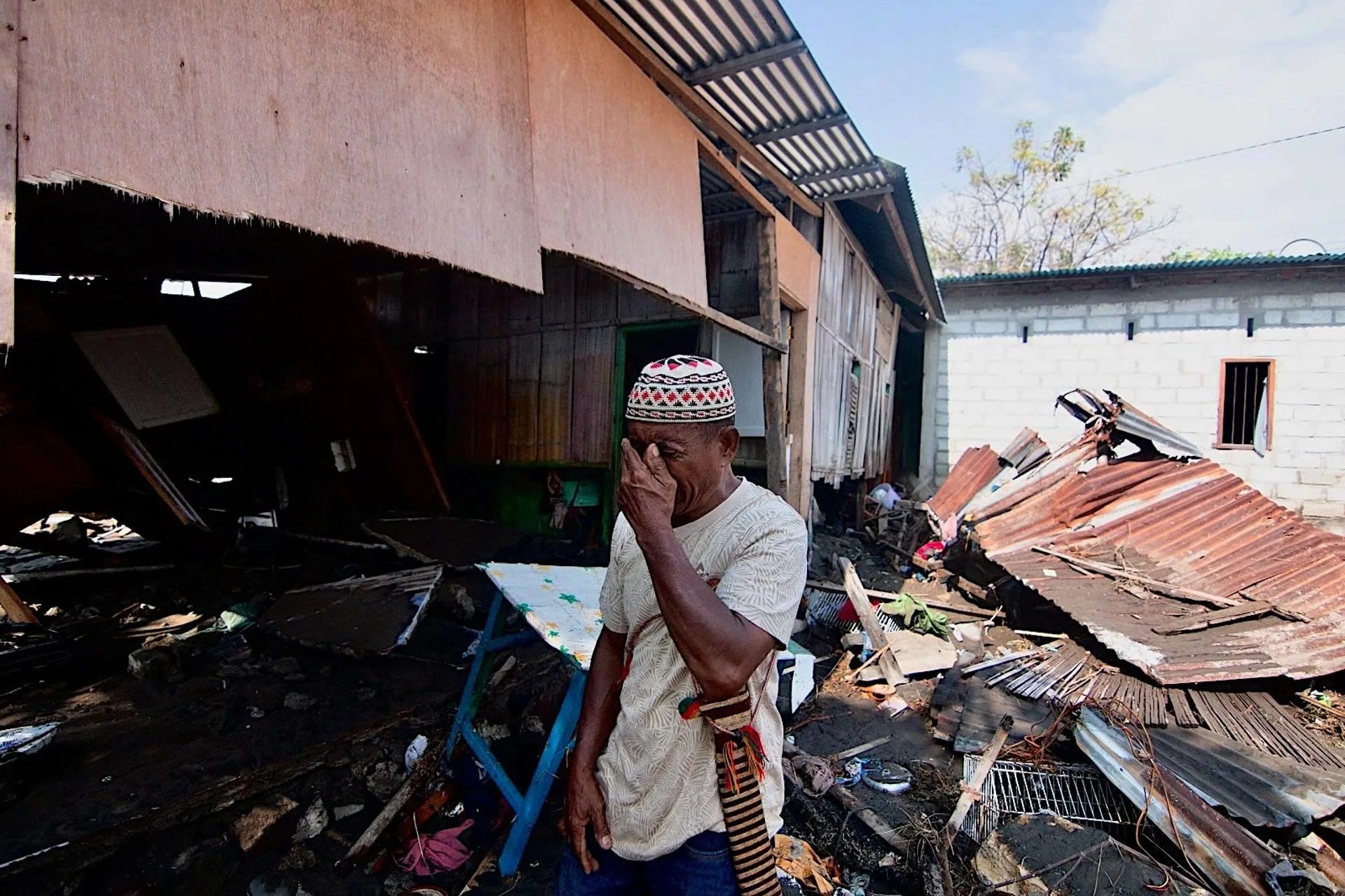 An older man in a white cap covers his eyes with his hand as he stands in front of a destroyed wooden house and a pile of rusted roofing.