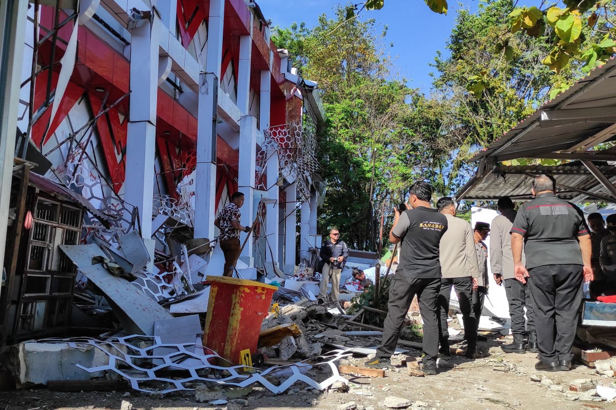 Men in uniforms stand among torn facade panels and debris outside a damaged red and white building.