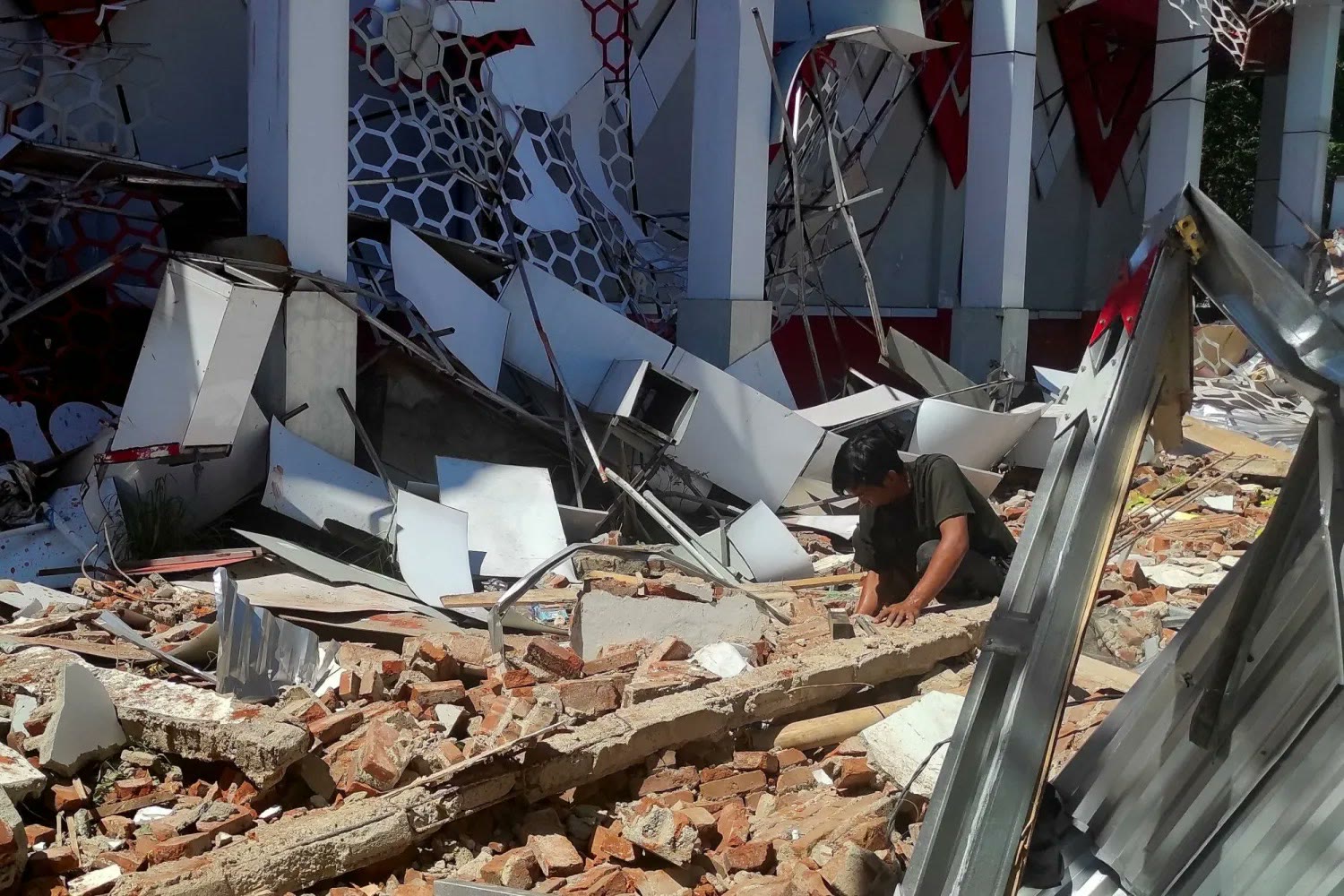 A man crouches in a field of broken bricks and twisted metal sheeting, searching through the rubble of a collapsed building.